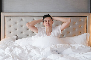 Young male sitting on bed in the morning after getting up. Handsome young man resting sitting in his bed. Young man in white t shirt feeling comfortable and sitting on bed in morning.