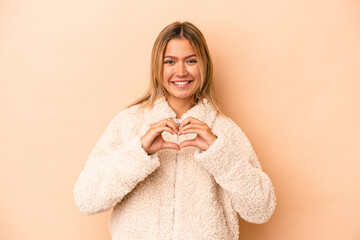 Wall Mural - Young caucasian woman isolated on beige background smiling and showing a heart shape with hands.