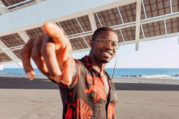 Cheerful black man with goatee style, listening music with earphones, pointing a finger outdoors under structure of solar panels at sunset in front of the sea. Smiling handsome african man.