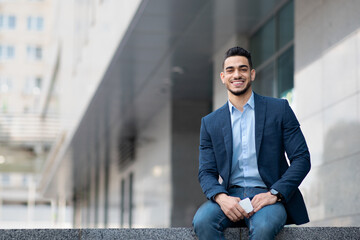 Cheerful middle eastern man in suit sitting by office building