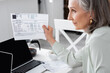 © LIGHTFIELD STUDIOS - Side view of blurred mature woman holding bills near laptops at home.