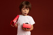 Portrait of upset Caucasian boy of 6-7 years old opening red present box, discontent with gift, having sad look, keeping mouth opened pronouncing sound of disappointment. Negative human emotions