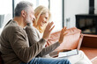 © Vadim Pastuh - Positive middle-aged couple is using laptop for video call sitting on comfortable sofa in cozy living room. Cheerful spouses waving into webcam. Online communication concept