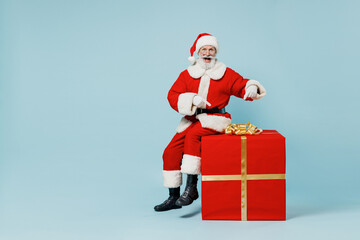 Full body old Santa Claus man in Christmas hat red suit clothes sitting point on big gift present box isolated on plain blue background studio. Happy New Year 2022 celebration merry ho x-mas concept.