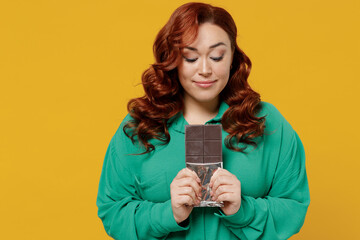 Excited happy hungry hungry smiling young ginger chubby overweight woman 20s years old wears green shirt hold in hand look down at chocolate bar isolated on plain yellow background studio portrait