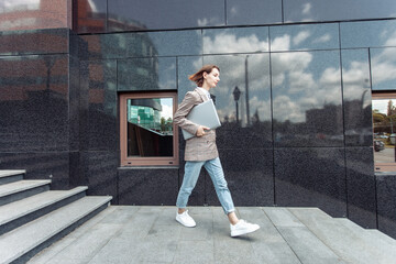 Wall Mural - Young informal business woman confidently walking with laptop in the city