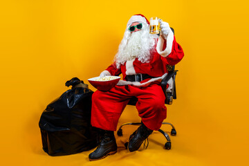 Santa Claus with a sack full of Christmas gifts is sitting for a break with popcorn and a beer isolated on orange background