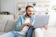 © Prostock-studio - Cheerful millennial man sitting on couch with laptop computer, working or studying online from home office