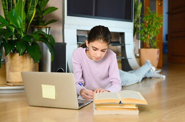 A girl at home on the floor with a computer and books is engaged in lessons.