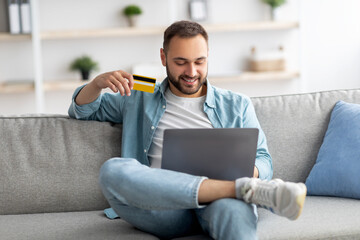 Wall Mural - Happy young Caucasian guy with laptop and credit card sitting on sofa at home, shopping online in web store