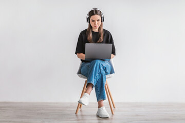 Serious young woman with laptop wearing headphones, sitting on chair, working or studying online, having video chat