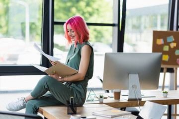 Wall Mural - cheerful businesswoman with pink hair and piercing holding folder and documents in office.