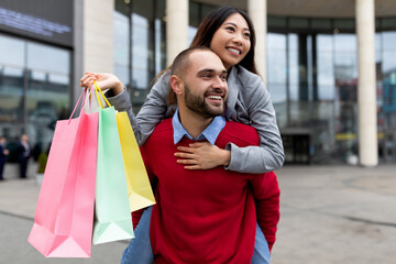 Wall Mural - Happy Caucasian guy giving his Asian girlfriend piggyback ride near shopping mall