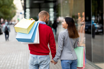 Wall Mural - Back view of young international couple walking hand in hand near big mall, holding shopper bags outdoors