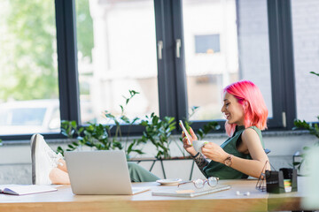 side view of happy businesswoman with pink hair holding smartphone and cup of coffee.