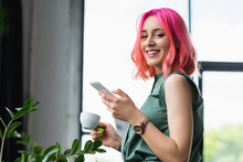 Cheerful Businesswoman With Pink Hair And Piercing Holding Cup Of Coffee And Smartphone.