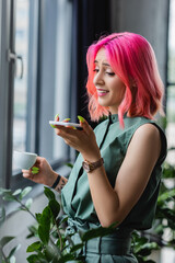 Wall Mural - cheerful businesswoman with pink hair and piercing holding cup of coffee while recording voice message on smartphone.