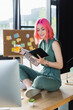 © LIGHTFIELD STUDIOS - happy businesswoman with pink hair holding cup of tea and looking at notebook while sitting on desk.
