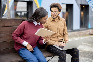 Black girl with digital tablet watching something on laptop with indian guy outdoors. Concept of remote and e-learning. Students lifestyle. Multiracial students sit on wood bench at university campus