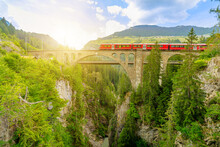 Red Train Crossing The Solis Viaduct Bridge Of Swiss Railway In Switzerland To Pontresina Town. Swiss Train Bernina In Grisons At Sunset. Albula Railway Section Between Thusis And Tiefencastel