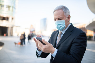 Wall Mural - Covid coronavirus concept, masked elder businessman using his smartphone outdoor