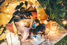 Father With Cute Children Draws Treasure Map Near Decorated Play Tent With Fairy Lights And Telescope Camping In Suburban Cottage Yard In Evening