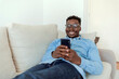 © Jelena Stanojkovic - Smiling African American man holding smart phone using social media apps sitting on couch at home. Black guy texting, remote learning, ordering distance delivery or communicating online in application