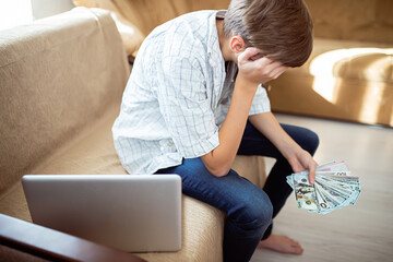 Sad teenager looking at bundle of dollars in hands while sitting by laptop at home. Problems with money deposits and savings among adolescents. Early financial independence 