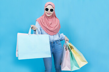 Portrait of excited happy young shopaholic beautiful asian muslim woman in traditional dress posing isolated over blue background holding pastel shopping bags and summer sale concept