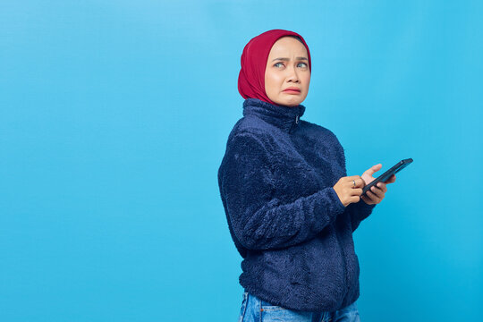 Scared young asian woman looking up and holding smartphone on blue background