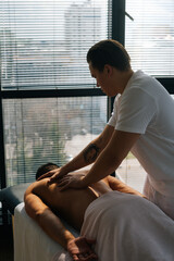 Wall Mural - Vertical back view of professional male masseur with strong tattooed hands massaging back and shoulders of muscular sports man lying on stomach, at massage table on background of window.