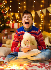 Wall Mural - Portrait of teenage boy in new year decoration. He eats sweets and cookies, has fun and a great mood. Holiday lights, gifts and christmas tree decorated with toys.