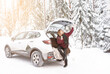 beautiful blonde young woman in a red sweater is sitting in the trunk of a car in the woods in winter among snow and trees