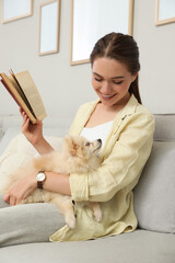 Sticker - Happy young woman reading book with cute dog on sofa in living room