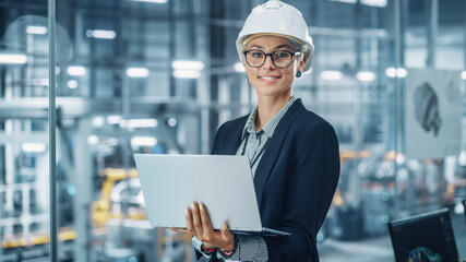 Leinwandbilder - Portrait of a Happy Young Beautiful Female Engineer Wearing White Hard Hat, Using Laptop Computer in Office at Car Assembly Plant. Industrial Specialist Working on Vehicle Design in Modern Facility.