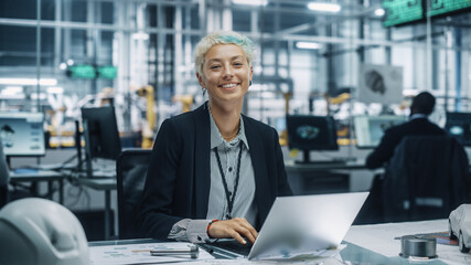 Wall Mural - Portrait of a Happy Young Beautiful Female Engineer Sitting at a Desk, Using Laptop Computer in Office at Car Assembly Plant. Industrial Specialist Working on Vehicle Design in Modern Facility.