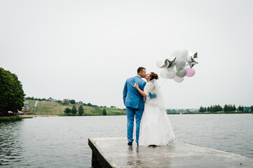 Wall Mural - Bride and groom hold white, grey, pink balloons near lake. Newlyweds kissing with helium balloons on ceremony. Couple in country. Wedding day. Back view.