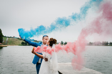 Wall Mural - Bride and groom holding blue and red colored smoke bombs near lake. Newlyweds with smoke bombs on beach.