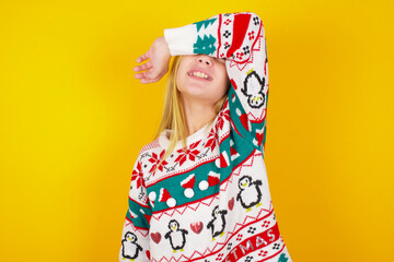 caucasian little kid girl wearing knitted sweater christmas over yellow background covering eyes with arm smiling cheerful and funny. Blind concept.