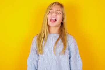 caucasian kid girl wearing blue knitted sweater over yellow background sticking tongue out happy with funny expression. Emotion concept.