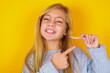 caucasian kid girl wearing blue knitted sweater over yellow background  holding an invisible aligner and pointing at it. Dental healthcare and confidence concept.