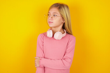 Wall Mural - Pleased caucasian little kid girl wearing long sleeve shirt over yellow background keeps hands crossed over chest looks happily aside