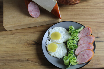 Canvas Print - breakfast traditional scrambled eggs fried eggs two ham sausages and cucumber salad on a gray plate on the table. next to it are products for cooking breakfast sausage bread. Horizontal photo