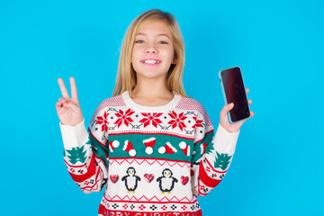  caucasian little kid girl wearing knitted sweater christmas over blue background holding modern device showing v-sign