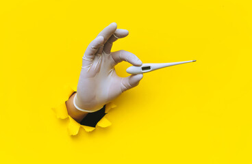 A right hand in a white medical latex glove is pushed through a torn hole in yellow paper and holds an electronic thermometer. The concept of diagnosing colds, covid-19, coronavirus. Copy space.