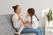 © sementsova321 - Extremely happy young adult mother playing with her charming daughter with pigtails pet a cake, expressing happiness, sitting on gray sofa in living room, laughing out loud.