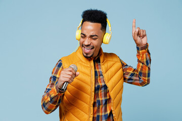 Wall Mural - Young black man 20s years old wear yellow waistcoat shirt listen music in headphones sing song in microphone have fun gesticulating hand isolated on plain pastel light blue background studio portrait