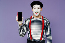 Fun Smiling Young Mime Man With White Face Mask Wears Striped Shirt Beret Hold Use Mobile Cell Phone With Blank Screen Workspace Area Isolated On Plain Pastel Light Violet Background Studio Portrait.