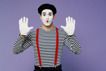 Wall Mural - Vivid young mime man with white face mask wears striped shirt beret looking camera hands raised as if touch stand near invisible wall isolated on plain pastel light violet background studio portrait.