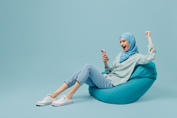 Full body young arabian asian muslim woman in abaya hijab sit in bag chair use mobile cell phone do winner gesture isolated on plain blue background People uae middle eastern islam religious concept.